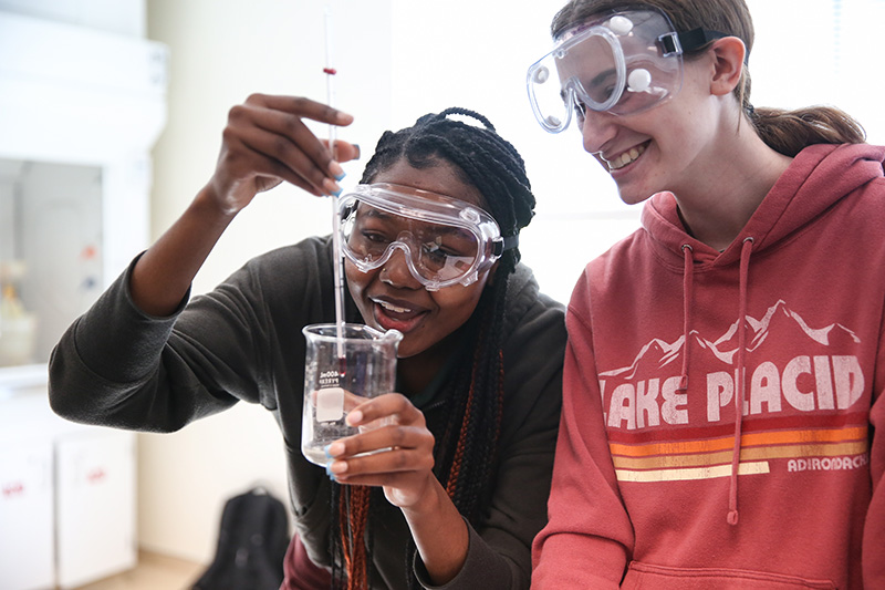 Two students wearing science lab goggles look at a substance in a chemistry beeker