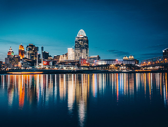 The downtown, Cincinnati skyline at nighttime. The lights of the buildings reflect off the Ohio River.
