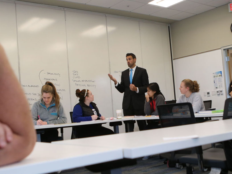 Students sitting at desks that are arranged to make a large rectangle. They are looking at a professor.