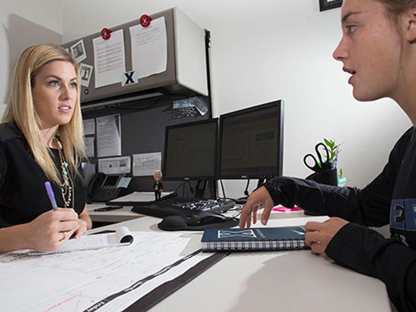 A student talking to an advisor. A desk is between them.