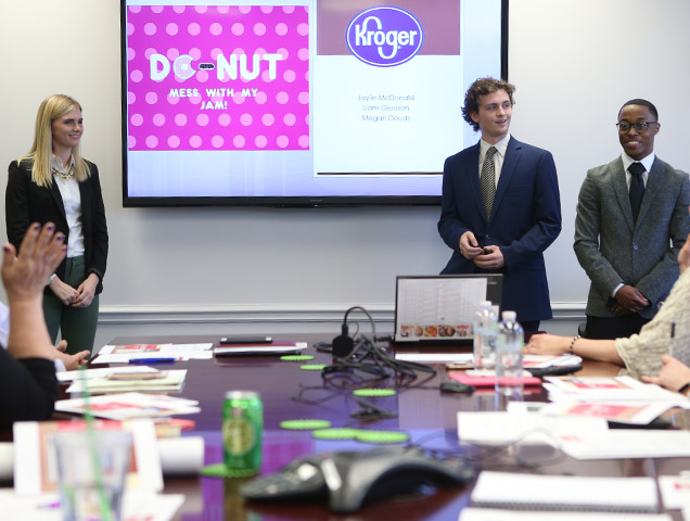 A group of students wearing business professional attire pitching an idea, projected on a screen behind them, to a room full of professionals 