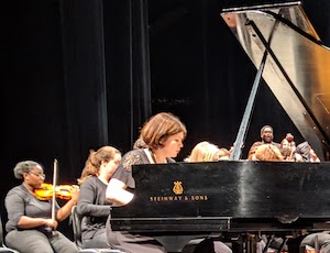 A Xavier professor sits behind a grand piano and plays the instrument. There is an orchestra of musicians behind her, including a violin player.
