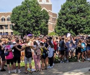 Students form two large circles by wrapping their arms around each other's shoulders. They are doing this as part of a new student orientation. They are outdoors in front of Gallagher Student Center. 