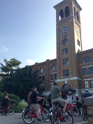 Three Xavier students riding red bicycles. They are stationary on their bikes. They are looking up at a clocktower on a building in Cincinnati.