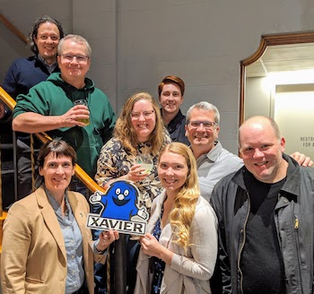 Eight members of the Xavier community stand together for a photo. They are all smiling. One person is holding a sign with an illustrated version of the Blue Blob (Xavier's secondary mascot).