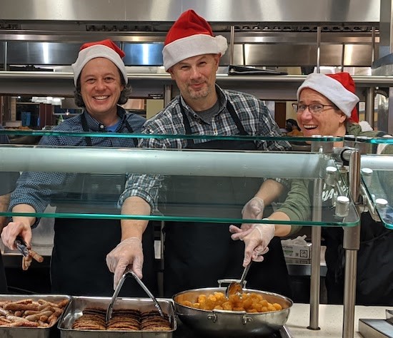 Three members of the Xavier community serve students food at the campus cafeteria. They are behind a hot counter. They are all wearing red Santa Claus hats.