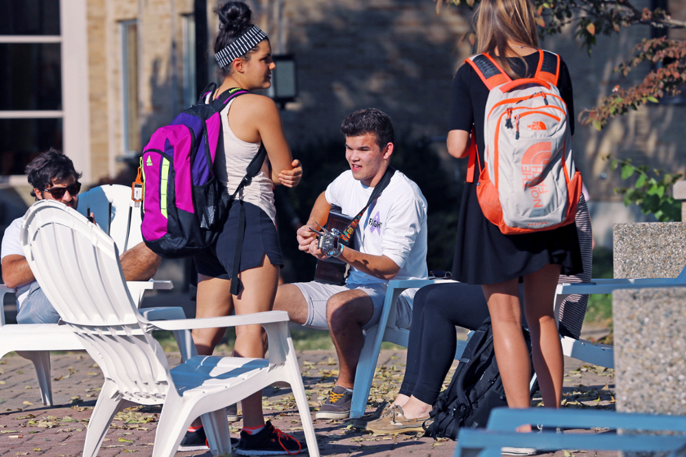 A student sits on a white Adirondack chair outside on Xavier's campus. The student is playing a guitar instrument for two students who are standing. 
