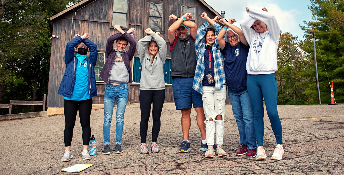 Group of seven volunteers with their arms in an X over their heads in front of a barn