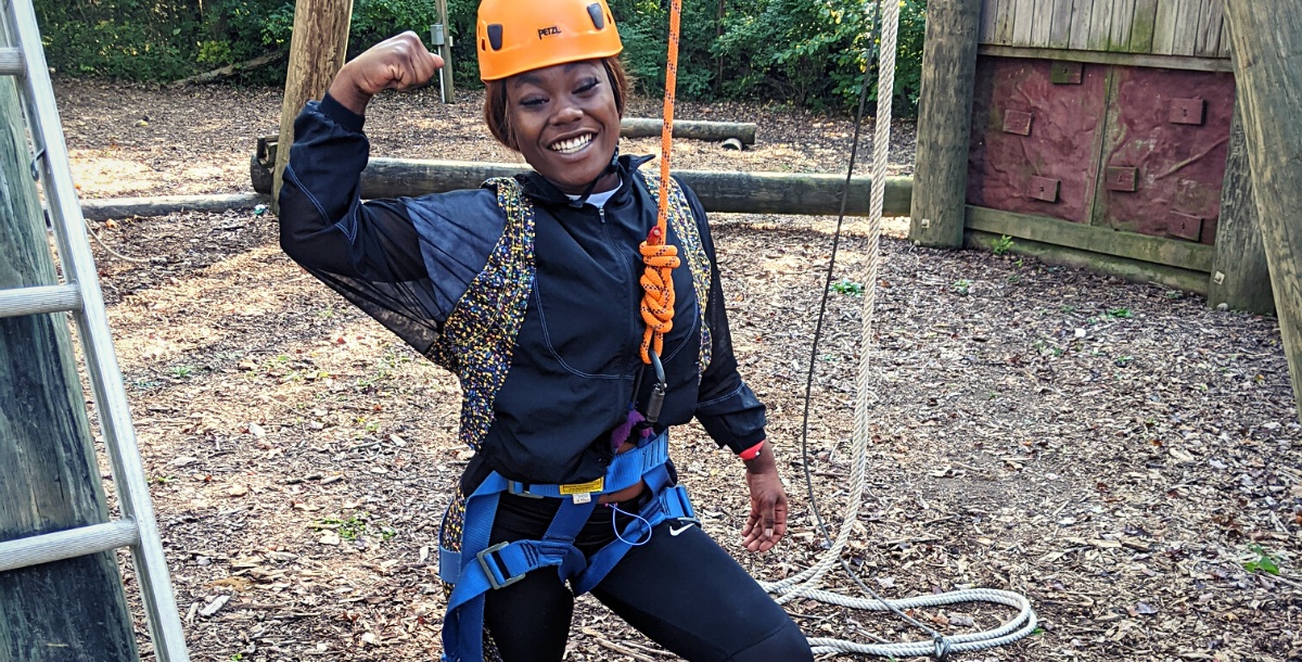 Student in a helmet flexing her biceps as she straps into a high ropes course