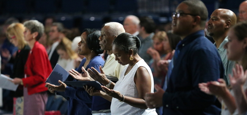 A row of parents standing during a mass. They have their arms out with their palms held upward.