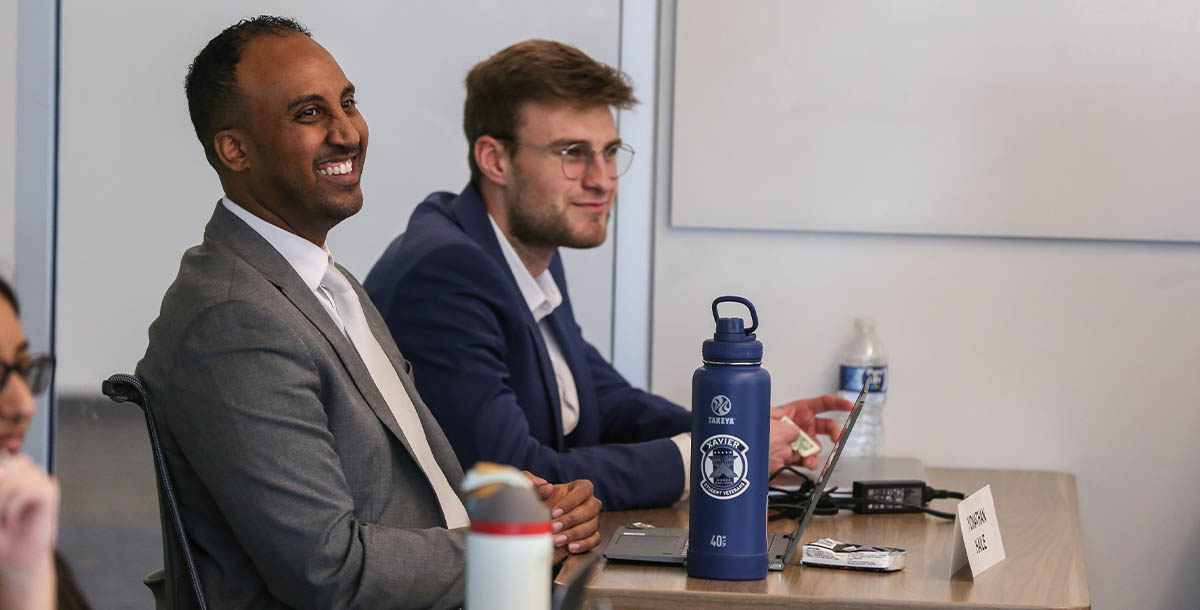 Two graduate students sit next to each other at a desk. They are smiling and wearing professional suits.