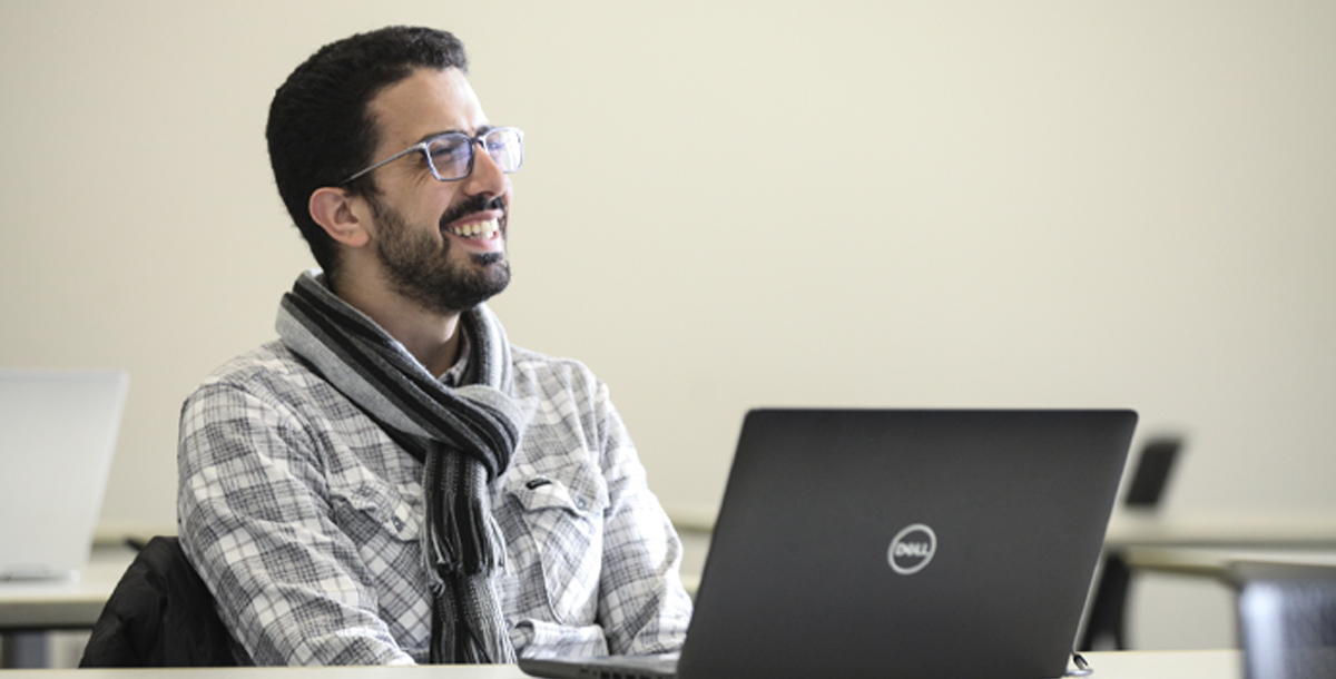 A student sits behind a laptop and smiles at someone off camera. The student is wearing glasses, a plaid button up shirt and a scarf.