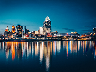 A photo of downtown Cincinnati, Ohio at night. Lights from the buildings reflect on the Ohio river.