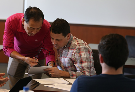 A professor and student are looking at a piece of paper together. The student is sitting and the professor is crouching beside the student.