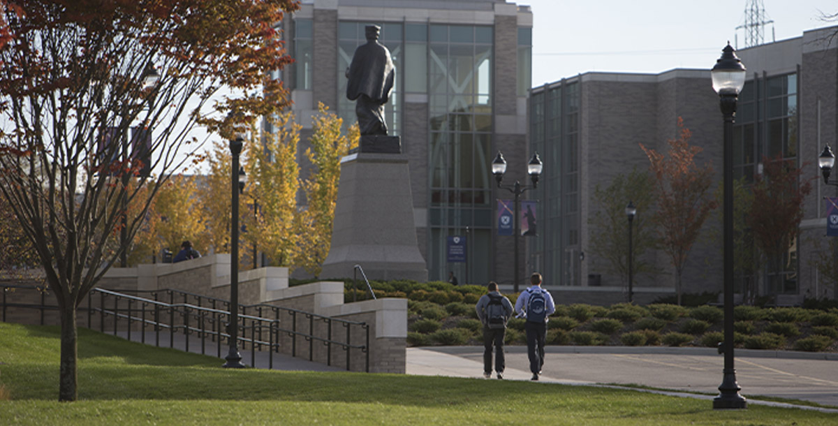 Two students walking along St. Francis Xavier Way at the Dana Avenue entrance to campus. It is fall and the leaves on the trees are beginning to turn.