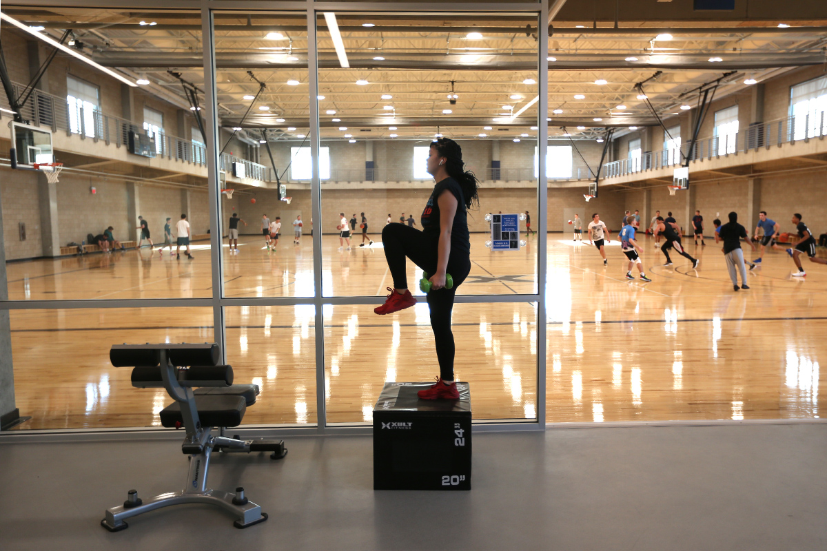 a student lifting arm weights in a room with glass walls.You can see the basketball court on the other side of the wall, where of a group people are playing basketball together.