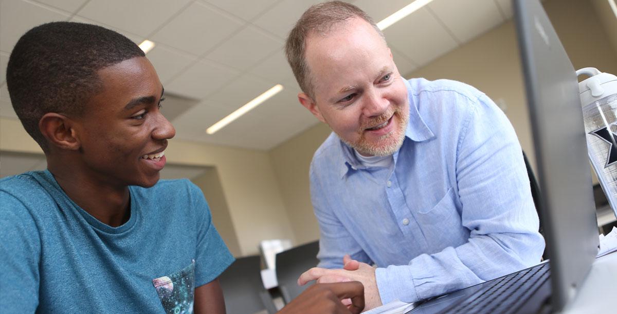 A Xavier professor and student sitting at a desk while looking at an open laptop screen.