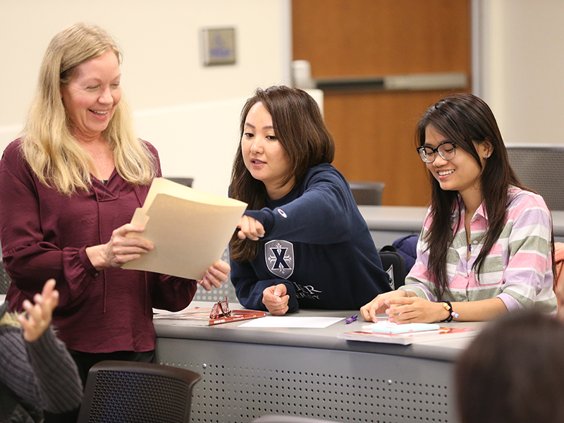 Two Xavier students sit at a long desk with laptops open in front of them. They are having a conversation with a professor who is standing to the left of them. The professor is holding a manilla file folder.