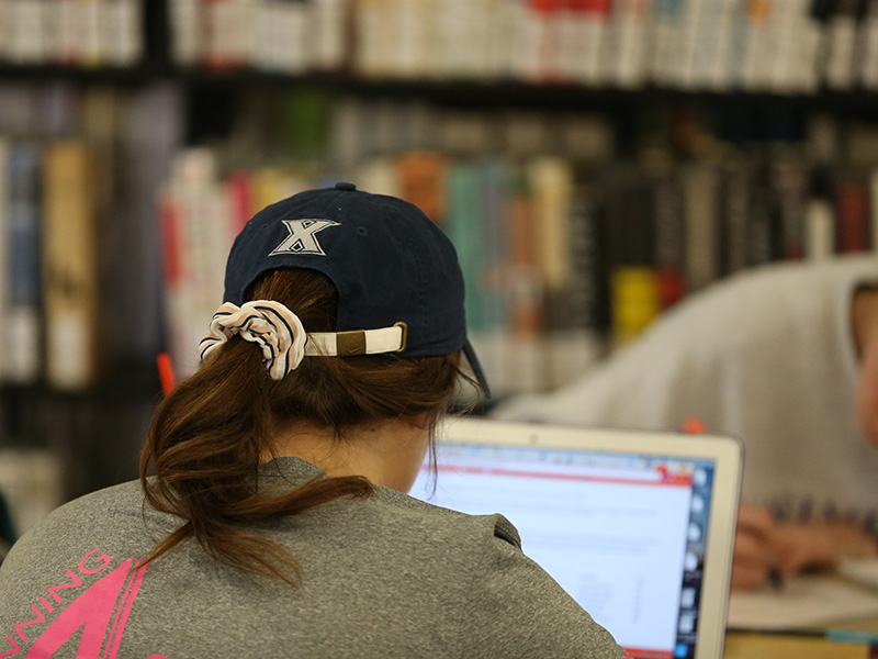A photo of the back of a students head. The student is sitting on a desk in a library. There is a laptop open in front of the student. There are shelves of books in the background.