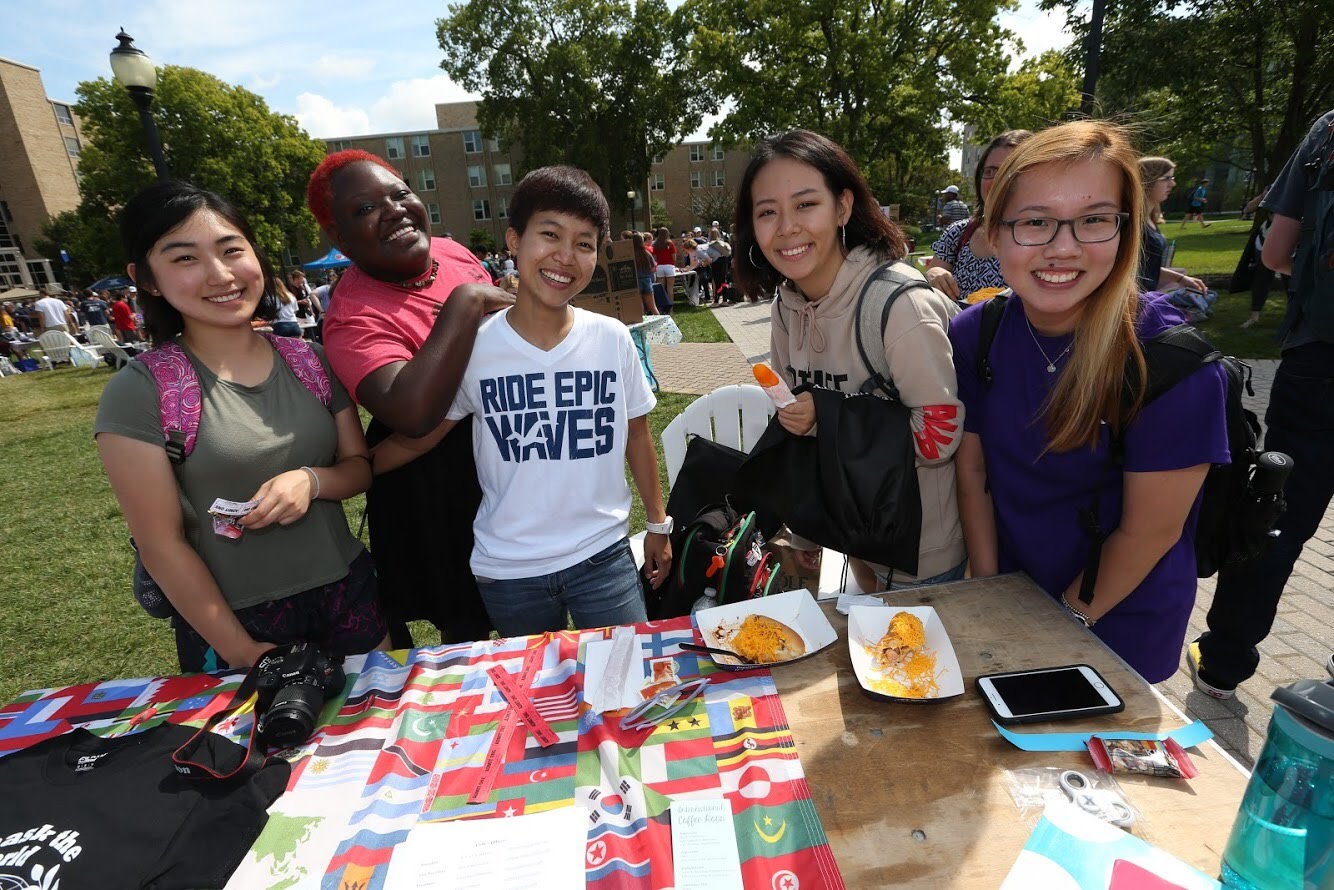 Four students smiling at their setup during Club Day on Xavier's campus. There are many different flags displayed on their table.