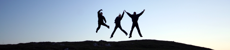 Silhouette of 3 people jumping gleefully into the air in front of a blue sky 