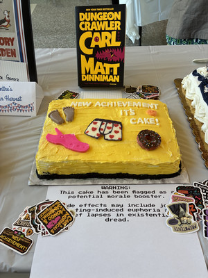 Photograph of a yellow cake with stickers surrounding it on a table. Book in background and paper with text in foreground.