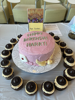 Photograph of a book, a pink cake with green writing on a stand surrounded by cupcakes on a table.