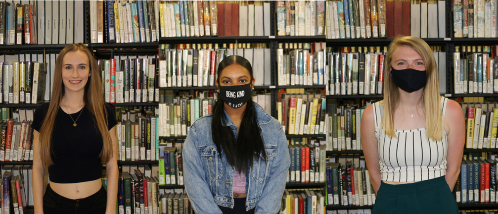 Nora Krukowski, Madison Martinez, and Emily Massey smiling in front of an aisle of books in the Xavier University Library