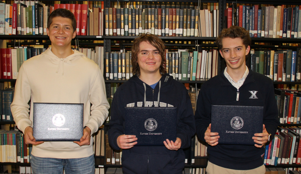 Luke Denecker, Aaron Ripley, and Sam Skelley holding their awards in front of an aisle of books in the Xavier University Library