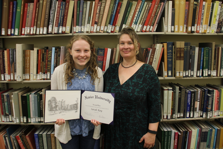 Sarah Ray holding her award smiling next to Dr. Renea Frey in an aisle of books in the Xavier University Library
