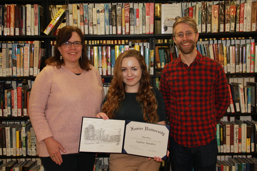 Assistant Library Director Alison Morgan, Sophie Boulter, and Dr. Timothy Brownlee smiling together in front of an aisle of books in the Xavier University Library