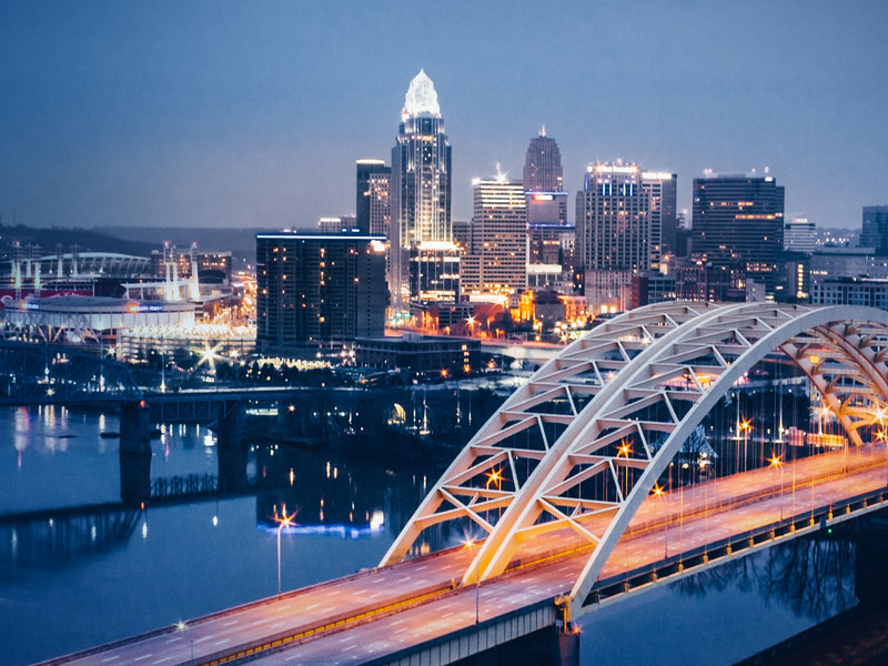 A photo of downtown Cincinnati at dusk, taken from Kentucky. The river and Roebling bridge is in the foreground.
