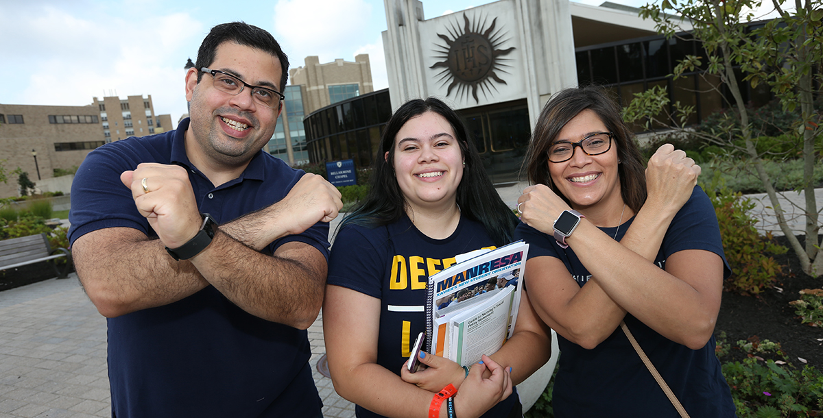 A Xavier student smiles with her parents in front of Bellarmine Chapel. All three of them are crossing their arms to form an X.