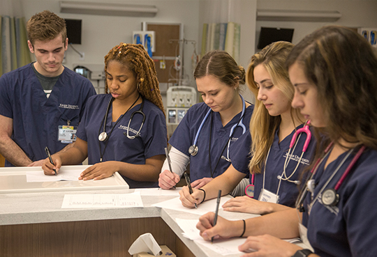Five Xavier nursing students in navy blue scrubs write on paper in a clinical setting