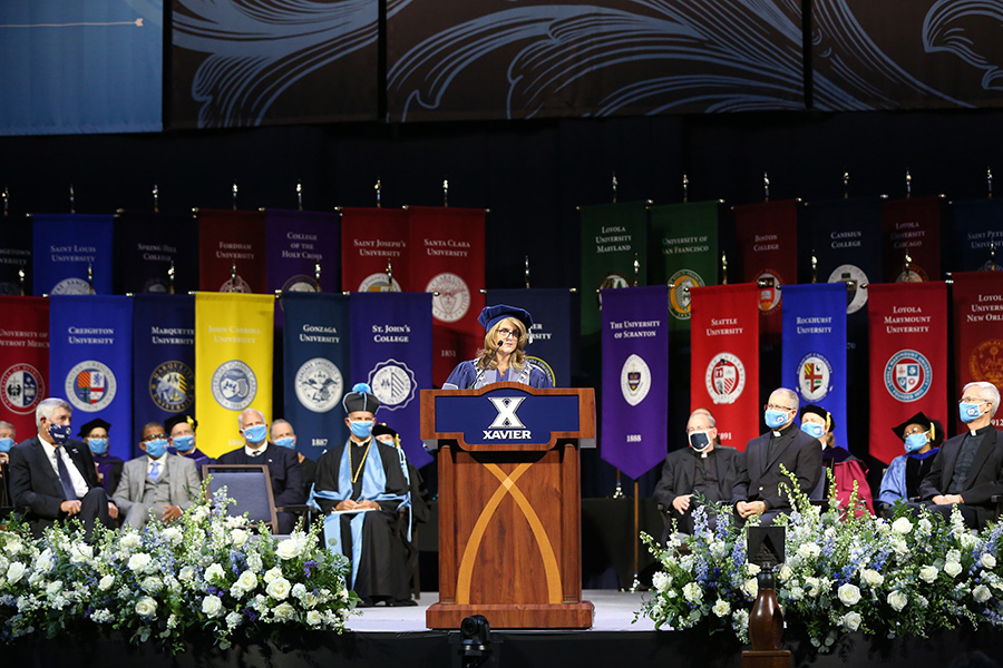 President Hanycz addressing the room during her installation ceremony.  She is standing behind a podium. There are flags representing each Jesuit university behind her. 
