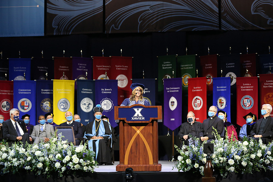 President Hanycz addressing the room during her installation ceremony. She is standing behind a podium. There are flags representing each Jesuit university behind her.