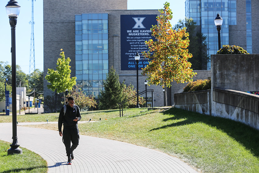 A man wearing a backpack walks through Xavier University's campus with a large banner containing the university's mission statement and several a large buildings standing in the background.