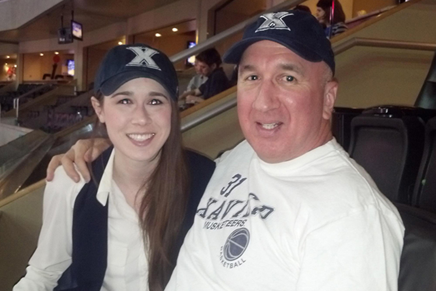 Emily Barbero and her father sit next to each other in the Cintas Center. They are both smiling and wearing Xavier baseball hats.