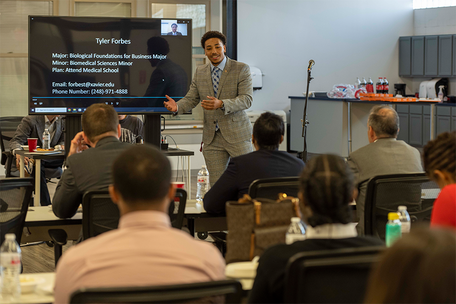 A student pitches their idea in front of judges. The student is standing in front of a screen.