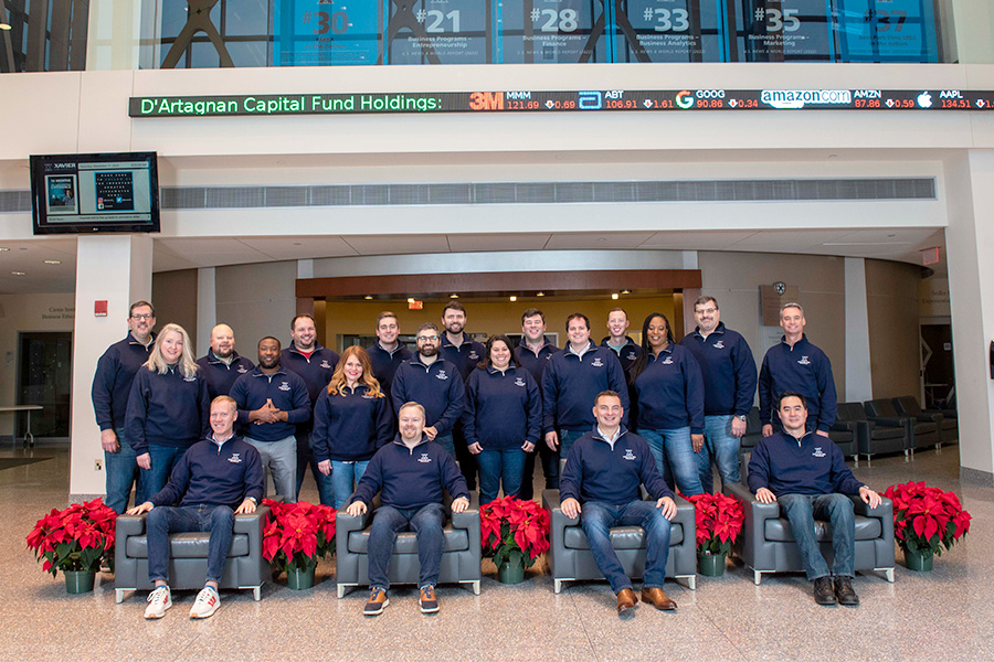 A group of Xavier University Executive MBA students pose in the atrium of the Williams College of Business with a stock symbol ticker overhead.