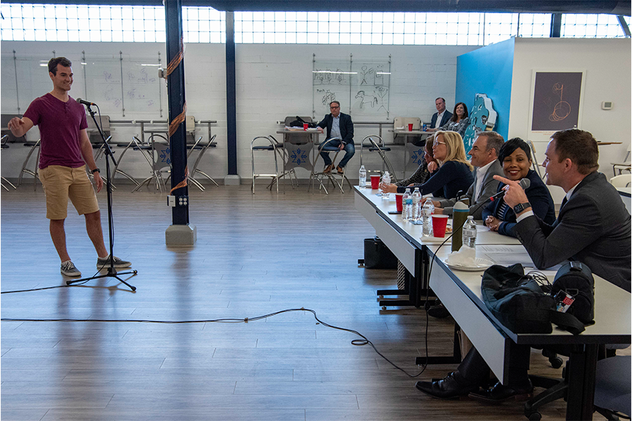 Xavier student pitches an idea in front of four TriHealth judges. The student is standing behind a microphone facing the judges, who are sitting behind a long table.