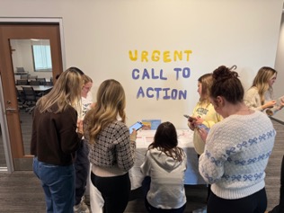 students kneel in front of booth with sign that reads 'urgent call to action'