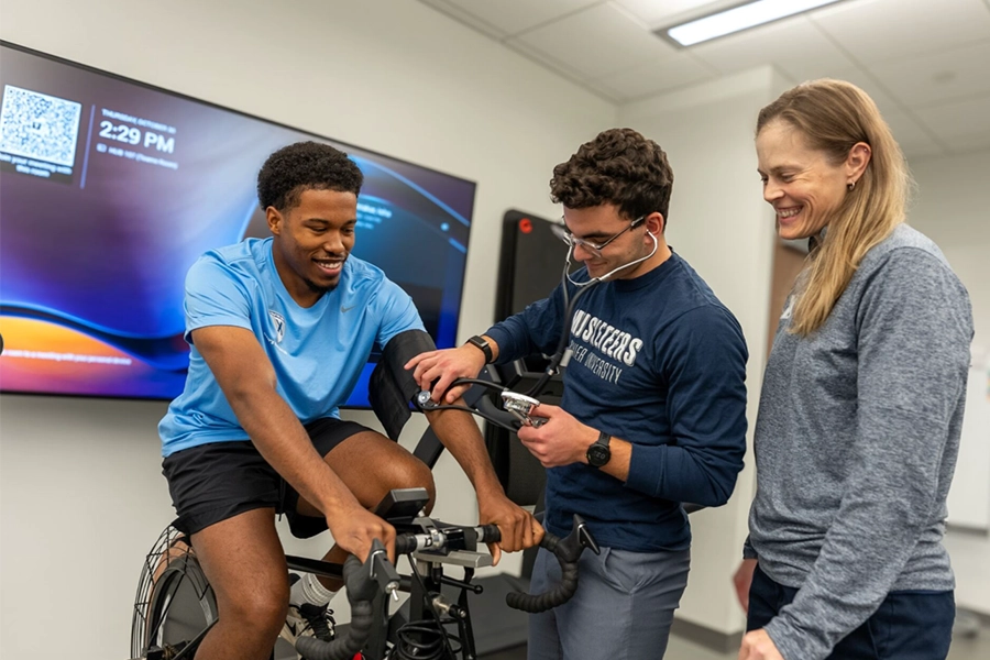 A student rides a stationary bike while another student takes their heart rate