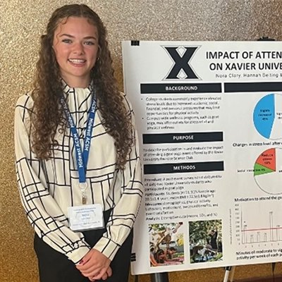 Nora standing in front of a research poster titled 'Impact of attending goat yoga on Xavier University student health'
