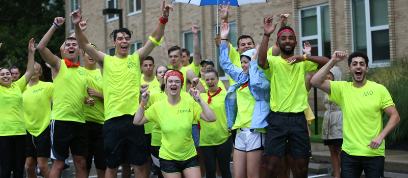 A group of 15 students wear neon shirts. They have their arms up in the air and are smiling. They are part of the Xavier Move Crew, upper-classmen who help first year students move to campus