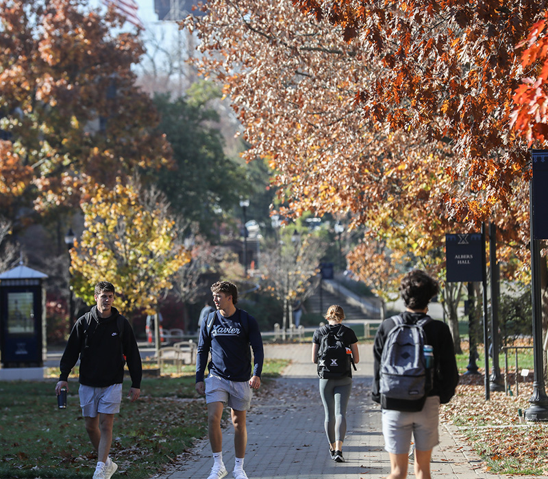 Four Xavier students walk down a paved sidewalk. They are surrounded by trees that are covered in red and orange leaves.