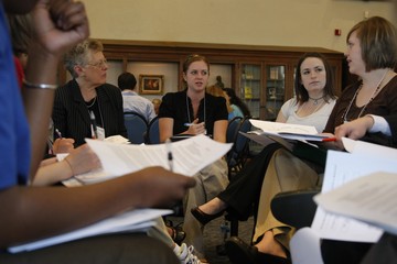 Five people sitting on chairs in a circle. They all have paper packets in their laps and are having a conversation.